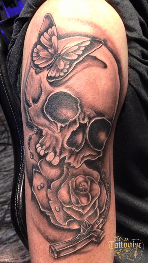 Zwart-grijze tatoeage op een bovenarm met een schedel, een vlinder, een roos en een kruis.