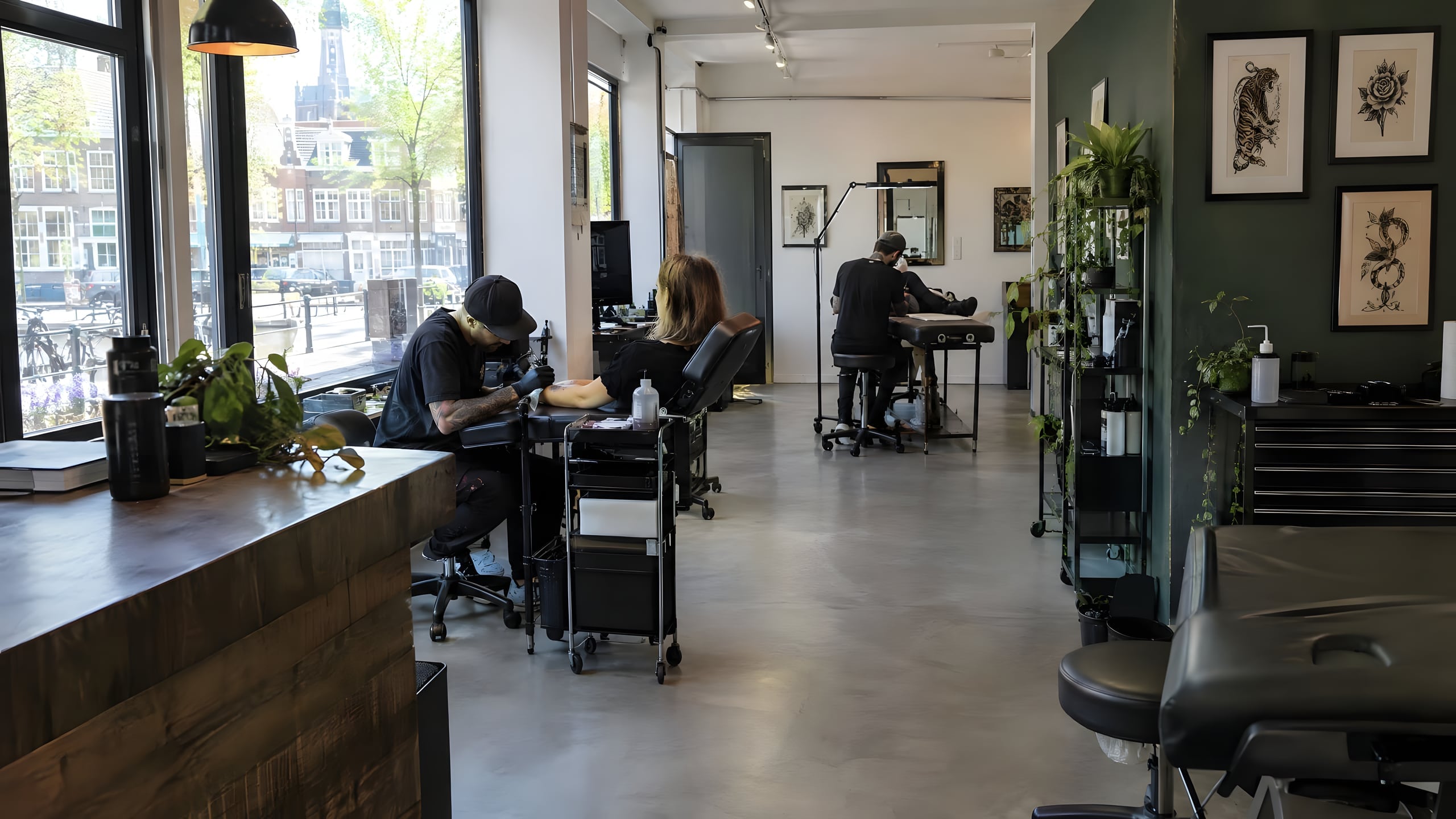 Tattoostudio met daglicht, een groene wand met ingelijst tekenwerk en twee tatoeëerders aan het werk.